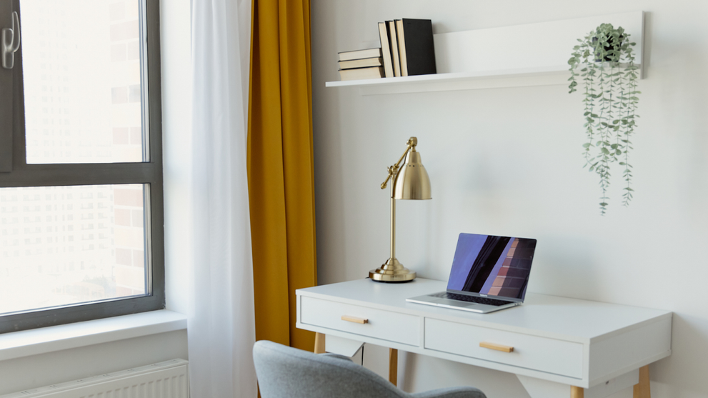 Minimalist home office setup with a white desk, gold lamp, gray chair, and laptop near a large window, featuring a wall shelf with books and a hanging plant